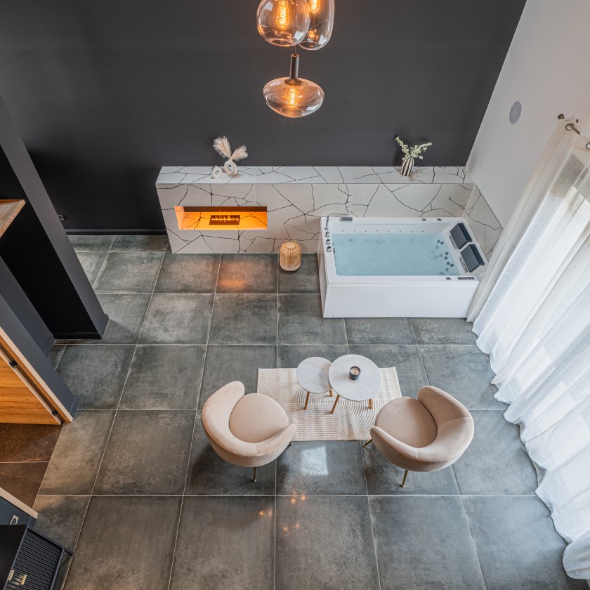 Espace de vie moderne avec jacuzzi intégré, cheminée encastrée et décoration épurée. Vue aérienne d’un salon lumineux avec fauteuils design et table en marbre, baigné de lumière naturelle à travers de grands rideaux blancs.